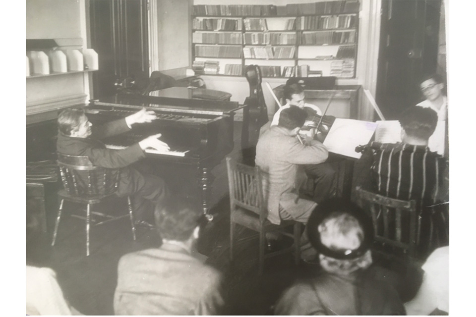 An old photo of string musicians performing in a circle, with a man at a piano raising his hands tot hem, with an audience watching behind them.